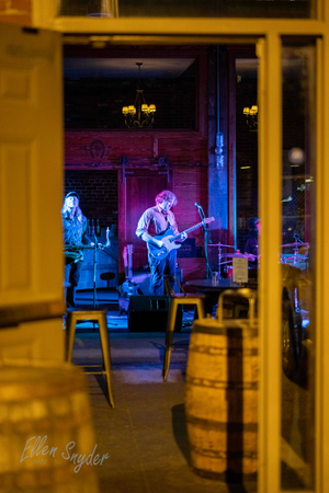Image is of musicians playing guitar as seen from the outside of the Boston Harbor Distillery door with whiskey barrels in front