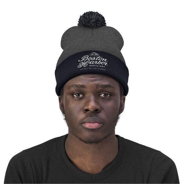 An image of a man wearing a Boston Harbor Distillery pom pom beanie in Navy/Graphite Heather