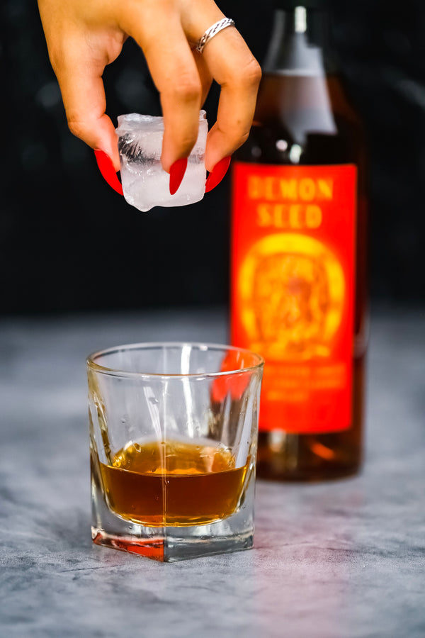 A rocks glass with Demon Seed and a hand dropping a large ice cube into the glass. Bottle of Demon Seed in background