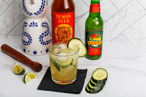 The Kickin' Mexican Mule cocktail with limes and cucumbers surrounding and bottles of tequila, Demon Seed and Reed's Ginger Beer in background