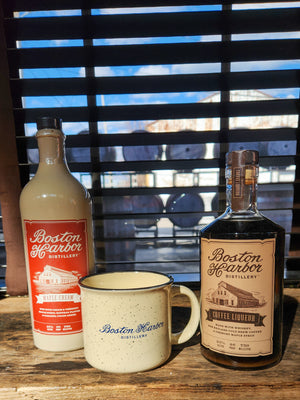 An image of a bottle of Boston Harbor Distillery Maple Cream and a bottle of Boston Harbor Distillery Coffee Liqueur on either side of a coffee mug sitting on a wooden window sill