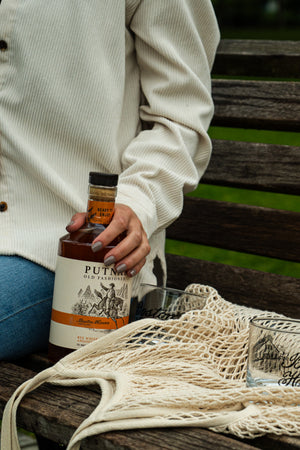 Image of a person seen only from the side with their hand on a bottle of Putnam Old Fashioned sitting on a bench with a bag and an empty glass