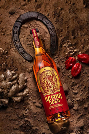A bottle of Demon Seed Whiskey laying on dirt with a black, metal horseshoe surrounding the neck of the bottle and 3 red Scorpion peppers on the right of the bottle and a bulb of ginger on the left of the bottle. Photograph by Igor Kragulac
