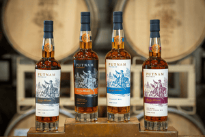 An image of four 750mL bottles of Putnam Whiskey varieties on a copper riser in front of a blurred background of whiskey barrels