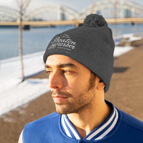 An image of a man wearing a Boston Harbor Distillery pom pom beanie in Graphite Heather